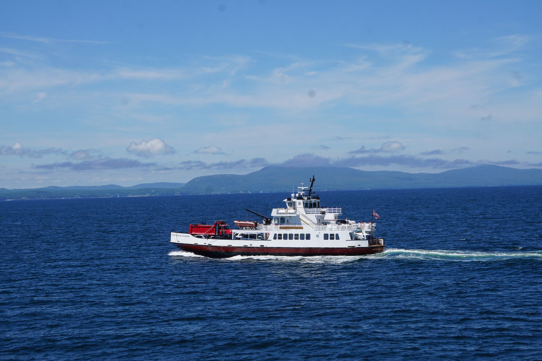 The Maine State Ferry vessel Captain Charles Philbrook sails toward Rockland with a load of vehicles and passengers. The foothills around Camden, Maine, dominate the skyline.